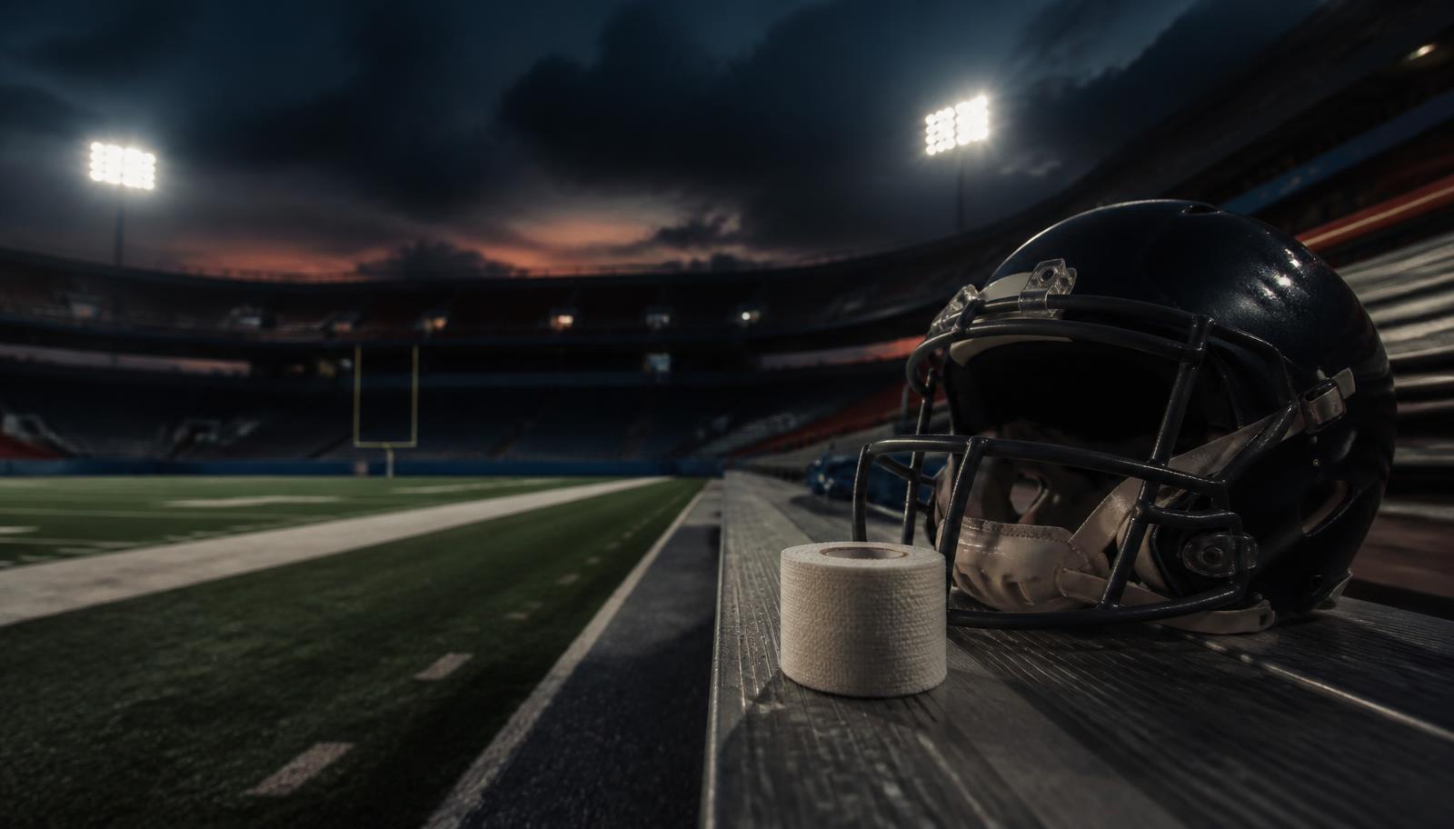 Empty NFL stadium at dusk with a helmet and a roll of athletic tape on the bench under floodlights.